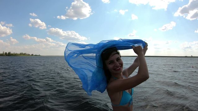 Slender girl with a big scarf on the shore. A woman holds in wind a developing scarf pareo stands near the river.