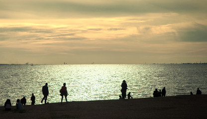Fototapeta premium Silhouettes of people walking along the shore of the sea bay against the sunset sky with clouds.