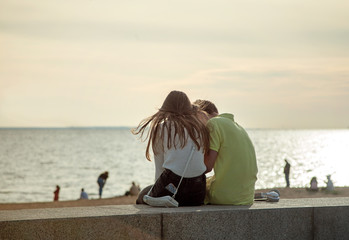 Enamored young girl and guy pair sitting on a granite parapet on the shore of the Gulf of Finland in St. Petersburg on a blurred background of a bay water and people on a beach.