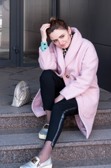 dreamy and smiling woman with bundled hair in pink coat and black pants sitting on stairs, straightens hair with hand and looking away near building