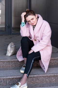 Dreamy And Smiling Woman With Bundled Hair In Pink Coat And Black Pants Sitting On Stairs, Straightens Hair With Hand And Looking Away Near Building