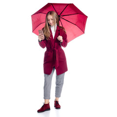 Woman in red coat standing with umbrella, smartphone mobile phone in hand looking autumn cold rain on white background isolation