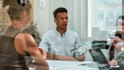 Business expert. Young afro american man holding eyeglasses and explaining something to his colleagues while sitting at the office table behind the glass wall in the modern office