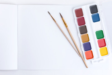 Top view on bright watercolor paints and two brushes for painting on an open album with empty pages for text or pictures on a white background