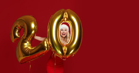 Portrait of young beautiful woman in fashion dress posing with gold 20 twenty number balloons isolated over red background. Christmas and new year Credit and deposit installments concept.