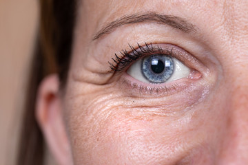 A close up and front view on the blue eye of a mature caucasian woman in her forties, showing heavy wrinkles (crow's feet) in the corner and below the eye, with copy-space. - obrazy, fototapety, plakaty