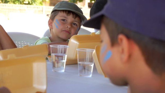 Group of children eating lunch from lunchboxes during the break between games and activities at school. The boy has a painted face
