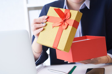 Christmas, any holiday. Office employee received a gift from her colleagues. Closeup woman hands holds gift box on office background.