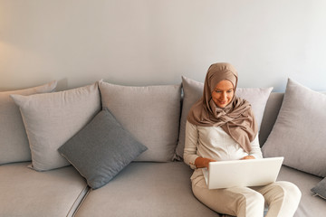 Muslim woman with laptop at home. Close Up of muslim business woman typing on laptop. Attractive young woman in head scraf using laptop while sitting on couch