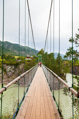 View of the road through a wooden suspension bridge with iron slings leading to a rocky island with a church over a green mountain river. The concept of a long road to a brighter future.