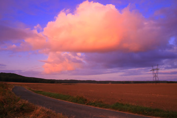 Big pink cumulonimbus in a blue sky over a field in the countryside. High voltage cable pylons.