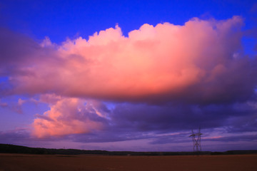 Big pink cumulonimbus in a blue sky over a field in the countryside. High voltage cable pylons.