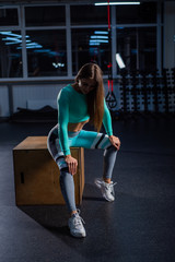 Fototapeta na wymiar A beautiful woman in a dark room sits on a crossfit cube. The coach is resting after a productive roundabout. Female athlete in sportswear in the gym.