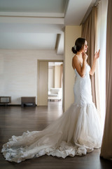 Side view of bride woman in wedding dress white looking trough the window from the room holding curtain