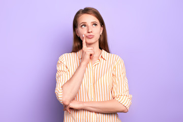 Dreamy young pretty female keeps index finger near lips, looking somewhere above, thinking what to wear on the first date, isolated over purple wall with blank space for your text.
