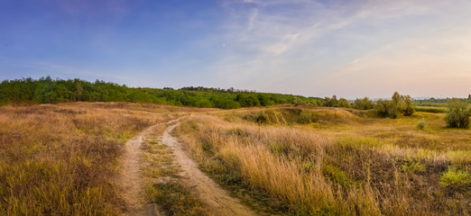 Idyllic autumn rural panorama with and a country track across a meadow with dry grass and hay. Beautiful evening scene, peaceful sunset light over the steppe vegetation.