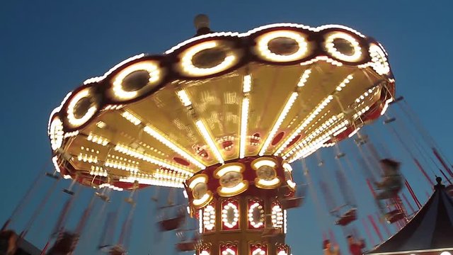 People ride at night on a carousel in the park. The carousel glows with different lights.