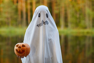 Waist up portrait of spooky child dressed as ghost holding pumpkin standing outdoors on Halloween,...