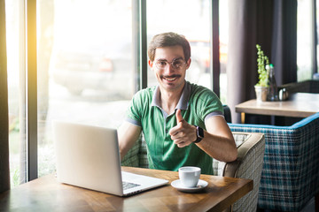 Like! Young happy businessman in green t-shirt sitting and working on laptop, looking at camera with thumbs up and toothy smile. business and freelancing concept. indoor shot near window at daytime.