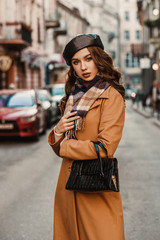 Outdoor autumn portrait of young elegant fashionable lady wearing trendy beret, camel color coat,...