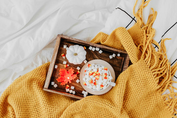 A cup of coffee with marshmallow and candy corn on bed with warm plaid. Autumn beverage with candle and pumpkin on wooden tray.   Hygge concept.