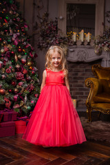 little blonde girl in evening red dress near the Christmas tree, child with Christmas gifts, the night before Christmas