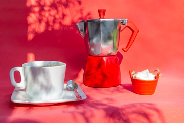 Italian coffee maker, moka pot coffee, cup and saucer of hot drink , sugar on red background with summer sunny day with shadow