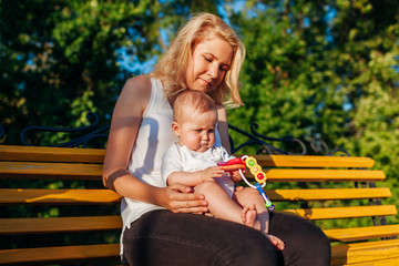 Baby girl playing with toys sitting on her mom's knees in summer park. Family having fun outdoors