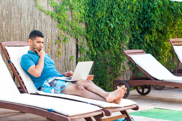 Portrait of clever handsome bearded young adult freelancer man in blue t-shirt and shorts lying on...