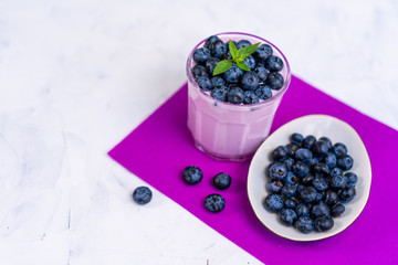 Tasty fresh blueberry yoghurt shake dessert in glass standing on white table purple napkin background.