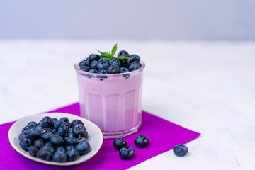 Fototapeta na wymiar Tasty fresh blueberry yoghurt shake dessert in glass standing on white table purple napkin background.