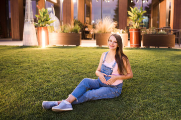 Smiling pregnant woman wearing denim  overalls sitting on green grass lawn outdoors in evening. Making heart on belly with hands. Looking at camera. Motherhood. Maternity.