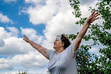 Senior woman practice yoga. She throws up her hands to the side. It is hard for her to do this. She's looking tensely straight. Old ages healthy lifestyle concept. Blue sky background. Low angle