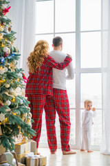 Happy family in plaid pajamas near the big window in the living room with a Christmas tree. Mother, father and little son waiting for Santa Claus