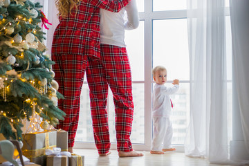 Happy family in plaid pajamas near the big window in the living room with a Christmas tree. Mother,...