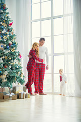 Happy family in plaid pajamas near the big window in the living room with a Christmas tree. Mother, father and little son waiting for Santa Claus