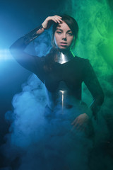 Obraz premium futuristic fashion model wearing black and silver clothes and standing in the colorful blue and green smoke