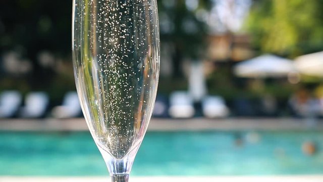 Close-up white champagne or prosecco glass against poolside at luxury resort hotel during vacation. Sparkling wine with rising bubbles with blue pool background outdoor. Refreshing alcohol drink