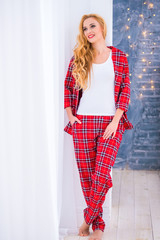 Beautiful blonde woman in red checkered pajamas stands near the window and demonstrates comfortable home clothes.