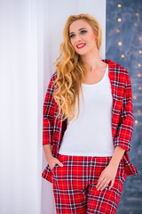 Beautiful blonde woman in red checkered pajamas stands near the window and demonstrates comfortable home clothes.