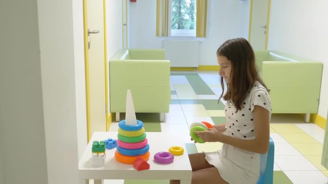 A girl sits in the lobby of a children's clinic, waiting for a meeting with a doctor, sorting a children's pyramid. The concept of medical examination, health monitoring, preventive examination of