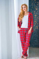 Beautiful blonde woman in red checkered pajamas stands near the window and demonstrates comfortable home clothes.