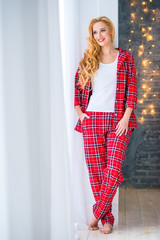 Beautiful blonde woman in red checkered pajamas stands near the window and demonstrates comfortable home clothes.