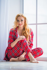 Beautiful blonde woman in red checkered pajamas sits near the window and demonstrates comfortable home clothes.