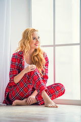 Beautiful blonde woman in red checkered pajamas sits near the window and demonstrates comfortable home clothes.