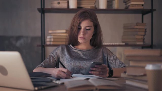 Young woman student in casual clothes studying at home table taking notes and using smartphone. Concept of modern technology in education. Handheld real time medium shot