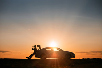 Silhouette of man driver relaxing after a ride, sitting on the trunk of his car and drinking water from a bottle, side view. Sunset time. 