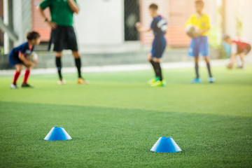 selective focus to blue marker cones are soccer training equipment on green artificial turf with blurry kid players training background. material for training class of football academy.