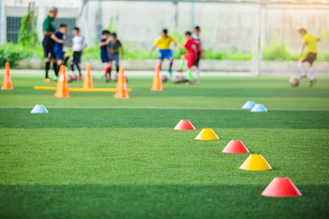 selective focus to red and yellow marker cones are soccer training equipment on green artificial turf with blurry kid players training background. material for training class of football academy.