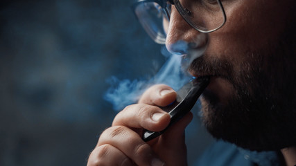 Man smokes new Vape Pod System, inhales and exhales vapor of electronic cigarette, vaping concept - obrazy, fototapety, plakaty